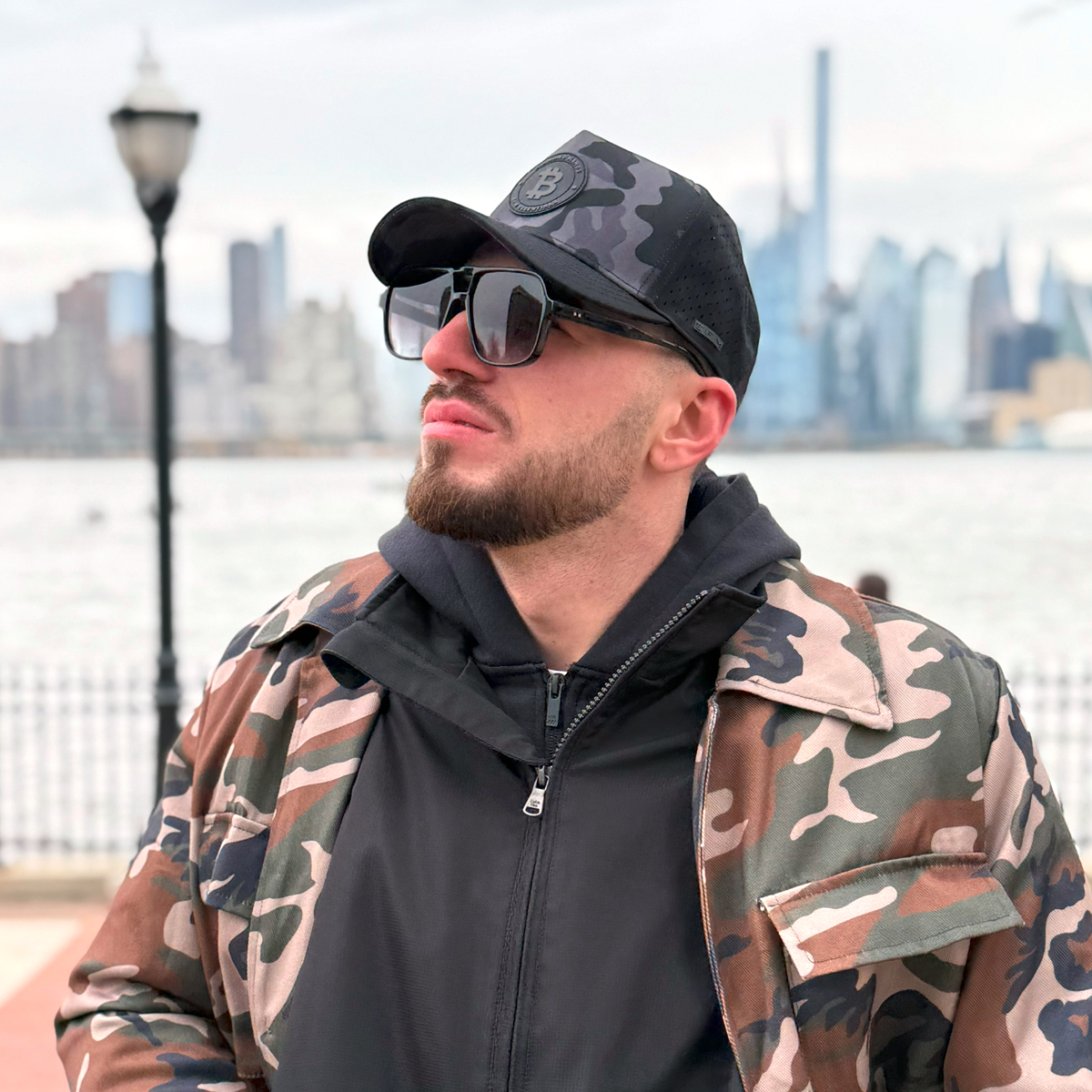 Man wearing a camouflage jacket and cap with sunglasses, standing outdoors with a cityscape in the background.