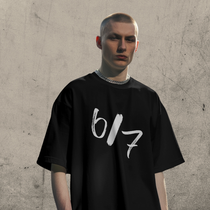 Person wearing a black t-shirt with '6/7' printed on it against a textured wall.