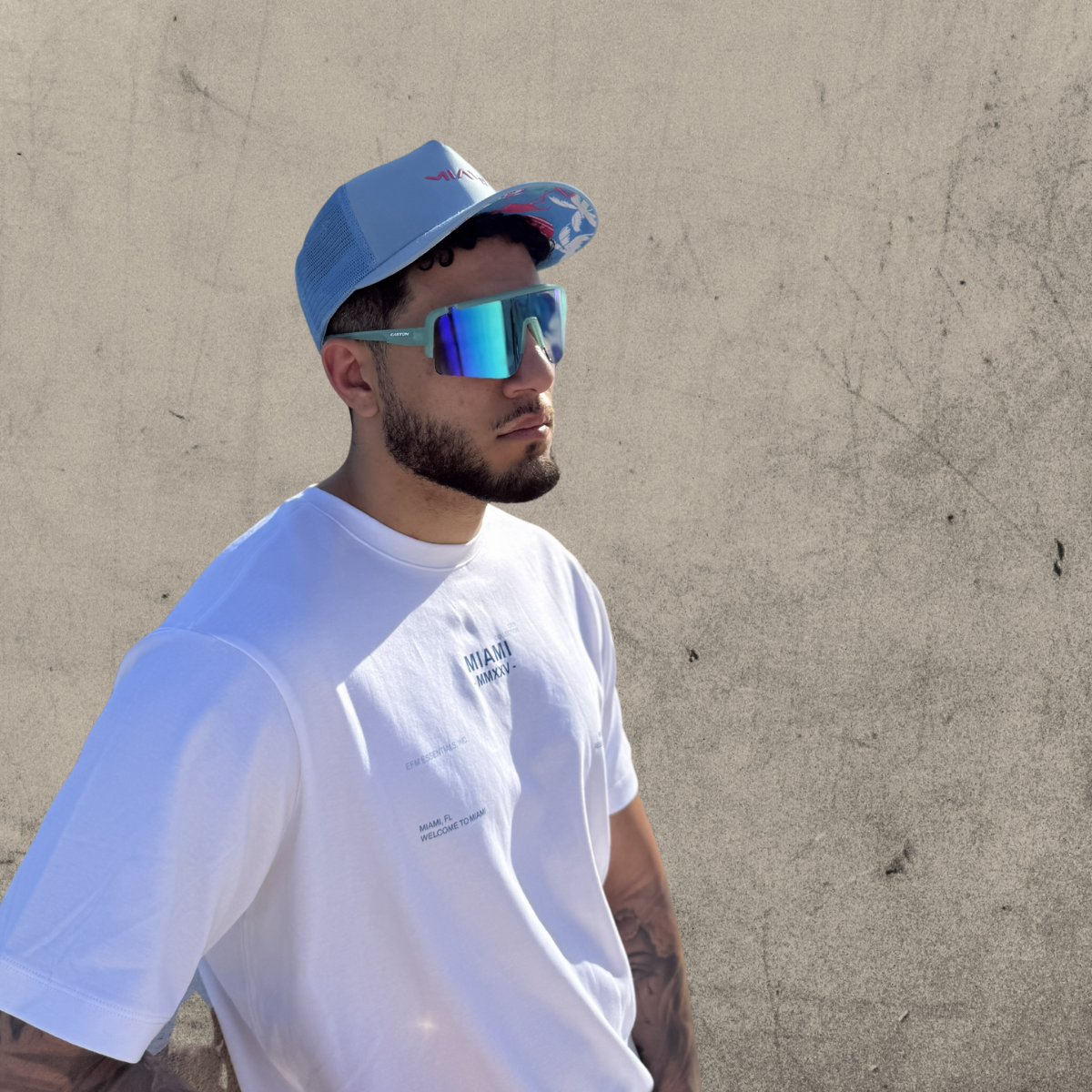 Man wearing a white t-shirt, blue cap, and reflective sunglasses against a beige wall.