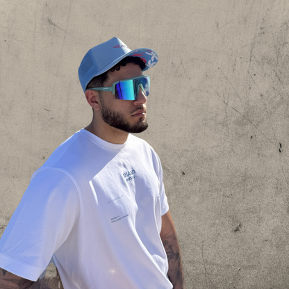 Man wearing a white t-shirt, blue cap, and reflective sunglasses against a beige wall.