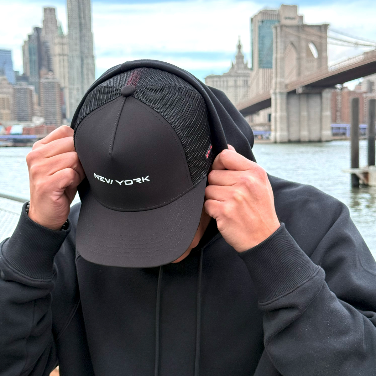 Person wearing a black cap with 'New York' text, standing in front of a cityscape with a bridge and water.